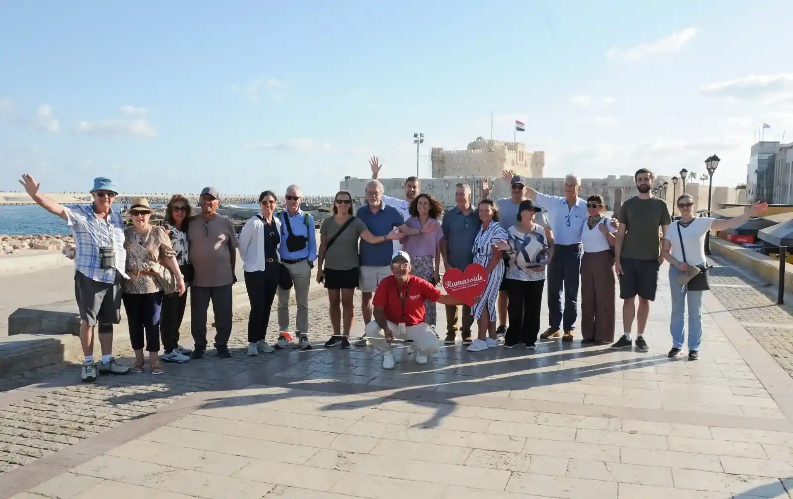 Happy travelers enjoying a sunny day trip in Alexandria, Egypt, standing near the Eastern Harbor with the 15th-century Qaitbay fortress visible in the distance.