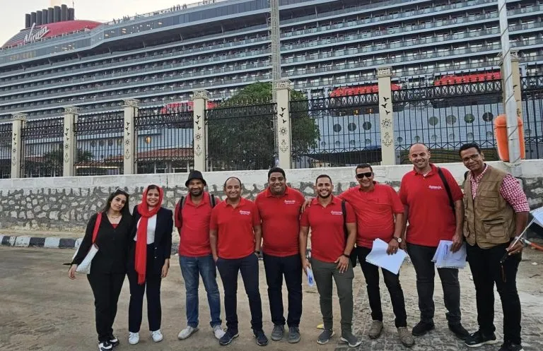 A group of Ramasside Travel Agency tour guides in red uniforms standing in front of a large cruise ship at Alexandria Port.